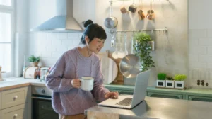 A woman holds a cup of coffee while using a laptop computer in the kitchen.