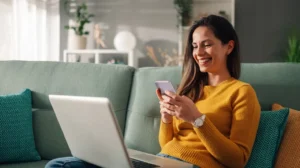 Woman in yellow shirt holding smartphone and smiling with laptop on her lap