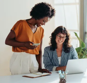 Two women smiling while looking at a laptop, discussing work.