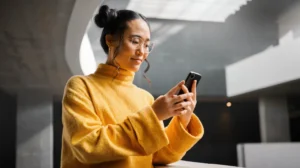 Woman wearing glasses and a yellow sweater smiling while using a smartphone indoors.