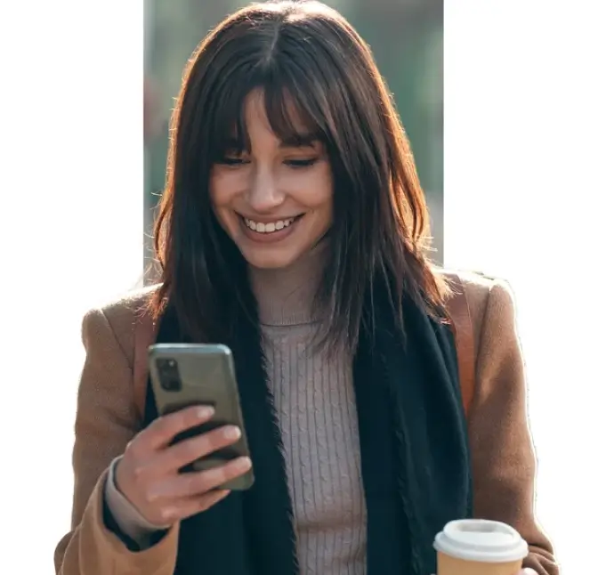 Smiling woman holding a coffee cup and looking at her smartphone while walking outdoors.