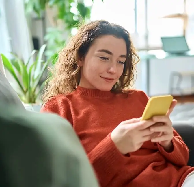 Woman sitting on a couch using a yellow smartphone in a bright living room.