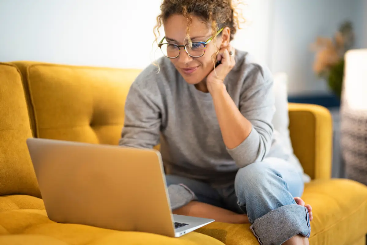 Woman sitting on a yellow couch using a laptop, focused on her screen in a bright and cozy living room.