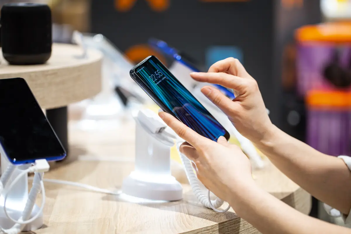 Hands holding and testing a smartphone on a display stand in a retail store.