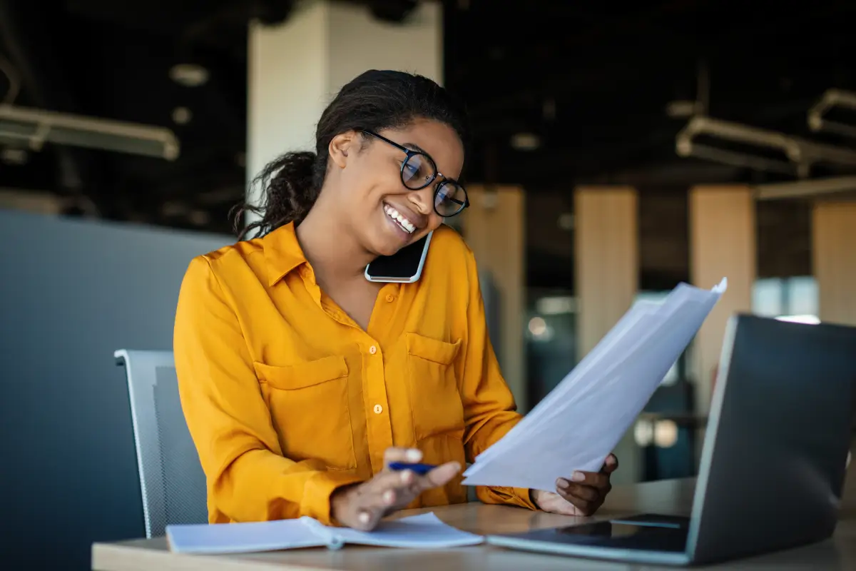 Smiling woman multitasking at her desk, talking on the phone while reviewing documents and working on a laptop.