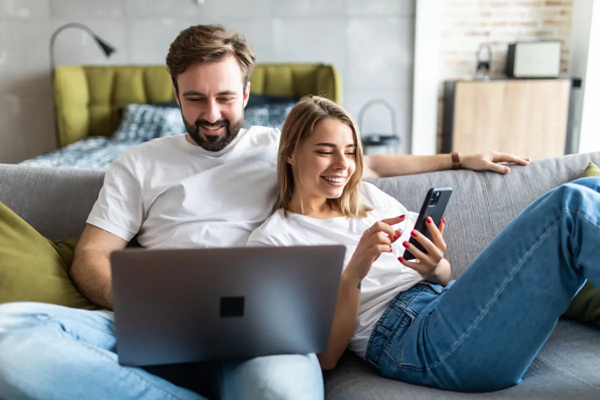 Happy couple relaxing on a couch using a laptop and smartphone together.