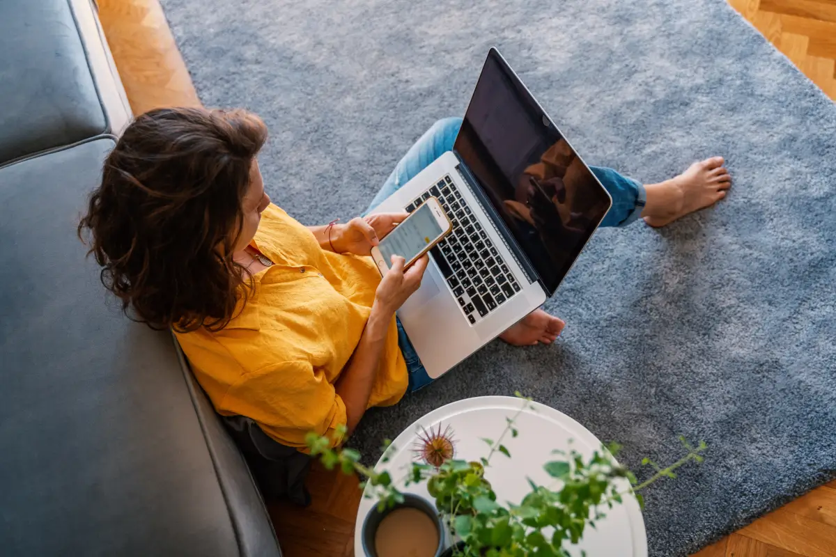 Woman sitting on the floor using a smartphone and laptop while working from home.