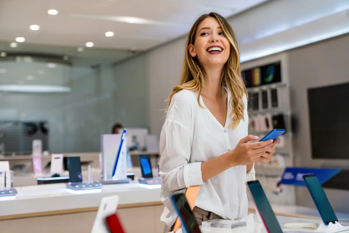 Smiling woman holding a smartphone inside an electronics retail store.