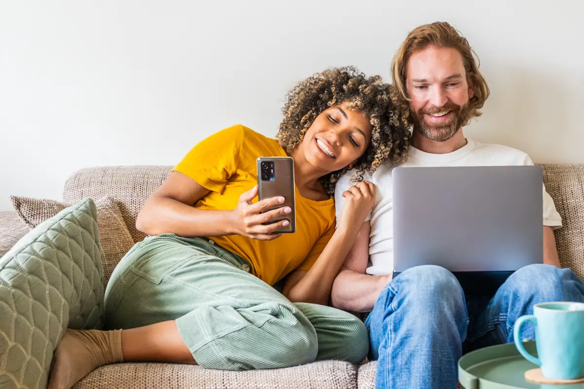 Couple relaxing on a couch using a smartphone and a laptop together.