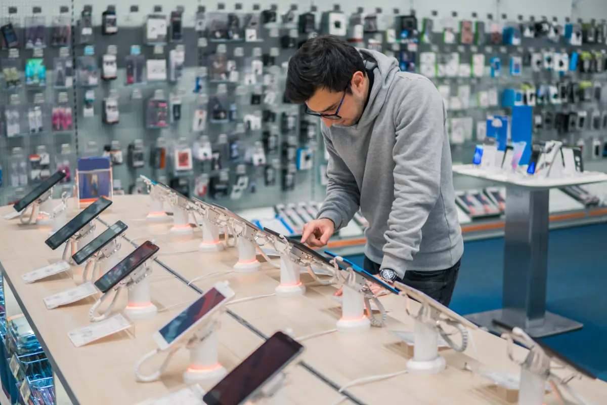 Man testing smartphones on display at a Verizon store with accessories hanging on the wall in the background.