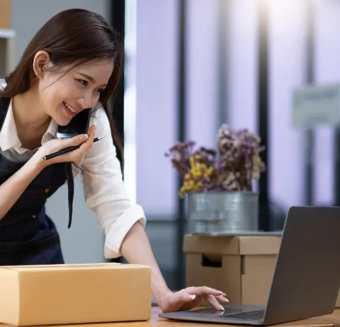 Woman smiling while talking on the phone and using a laptop next to a cardboard shipping box in a small business workspace.